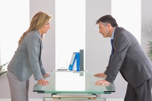 Two irritated businesspeople standing arguing on each side of a desk at office