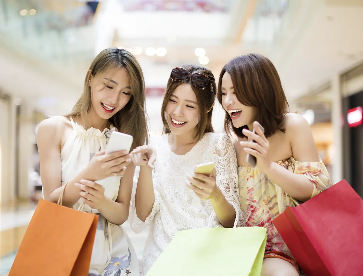 3 mujeres riendose con algunas compras en sus brazos revisando sus smartphones