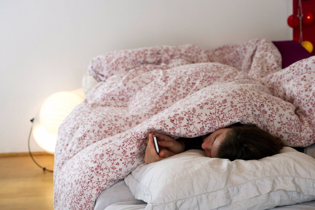 WOMAN LYING IN BED UNDER COVERS, LOOKING AT SMARTPHONE