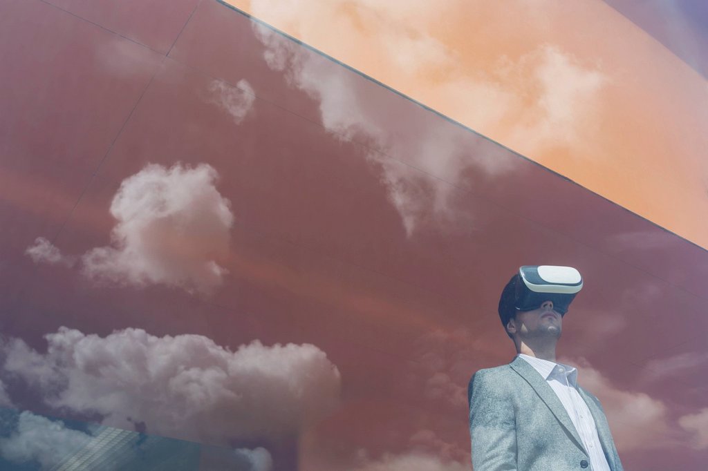 BUSINESSMAN USING VIRTUAL REALITY SIMULATOR GLASSES AT WINDOW WITH REFLECTION OF CLOUDS