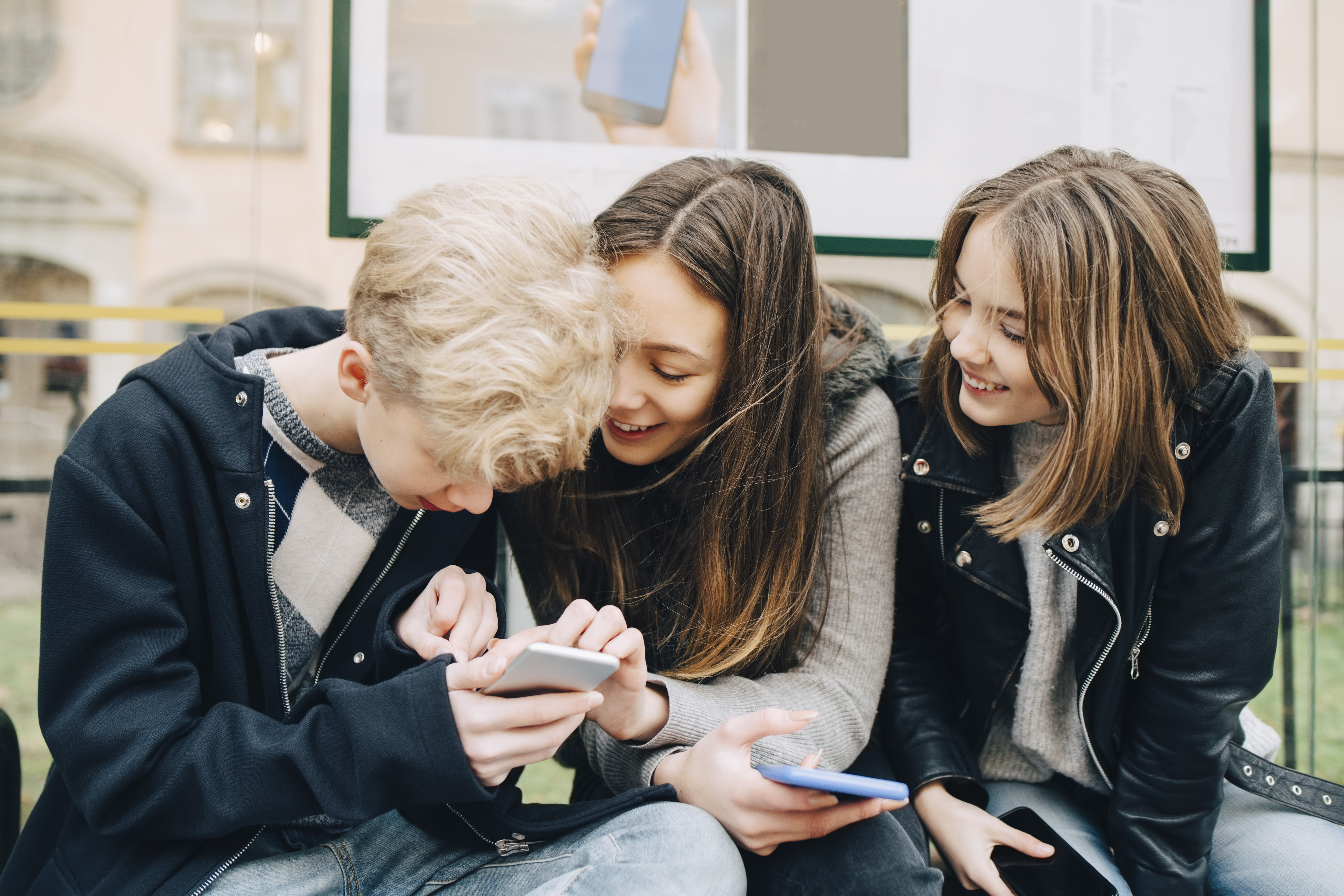 Smiling friends using mobile phone while sitting at bus stop. SuperStock 4448r-21966252