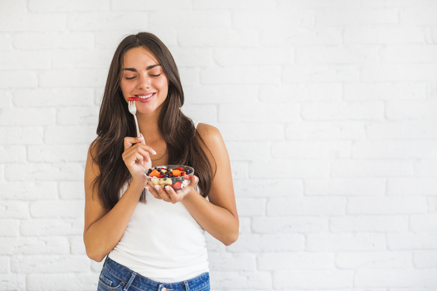 happy-young-woman-standing-against-white-wall-eating-fruit-salad