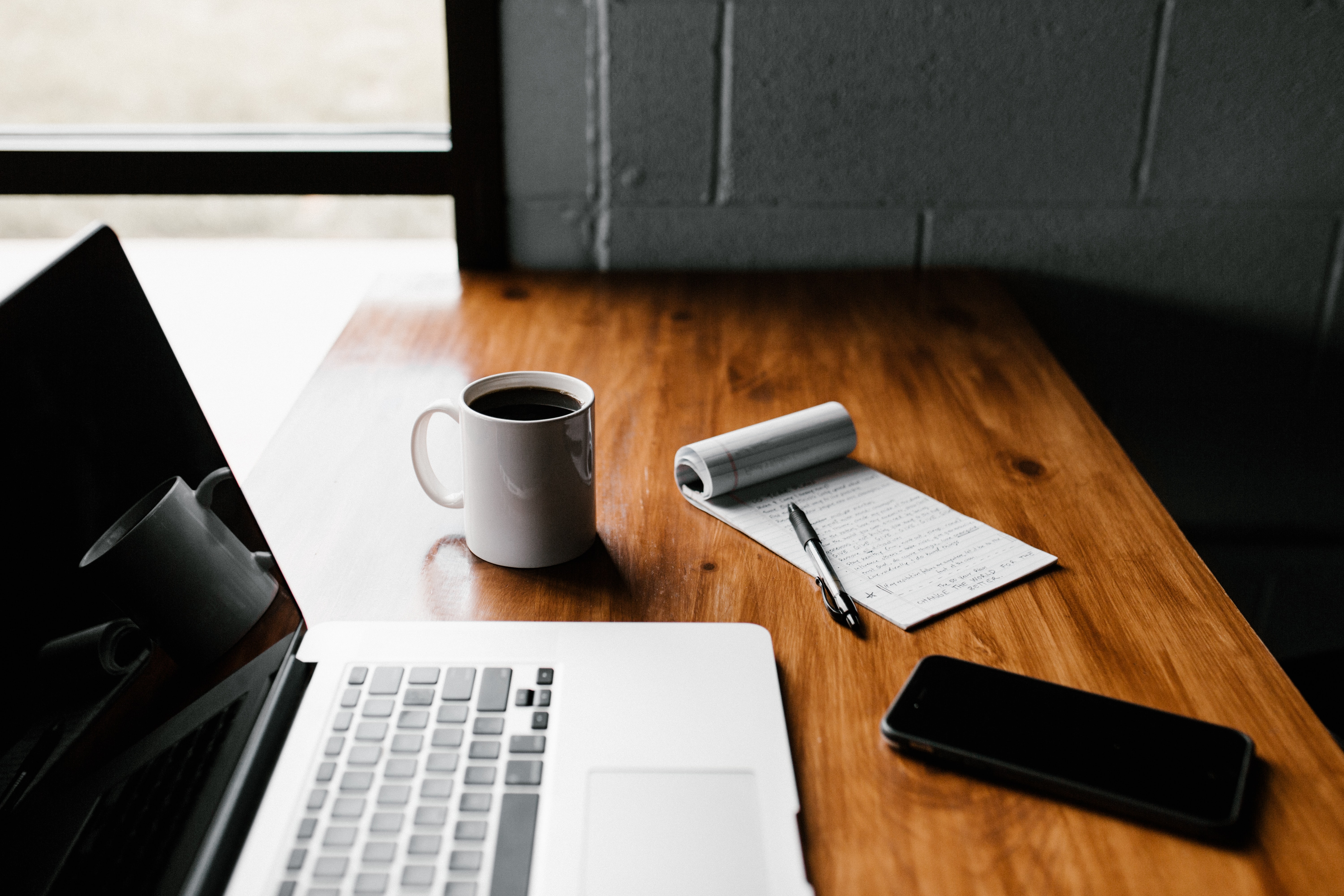 image of a pad and paper with a pen resting on top next to a computer, cell phone, and cup of coffee.