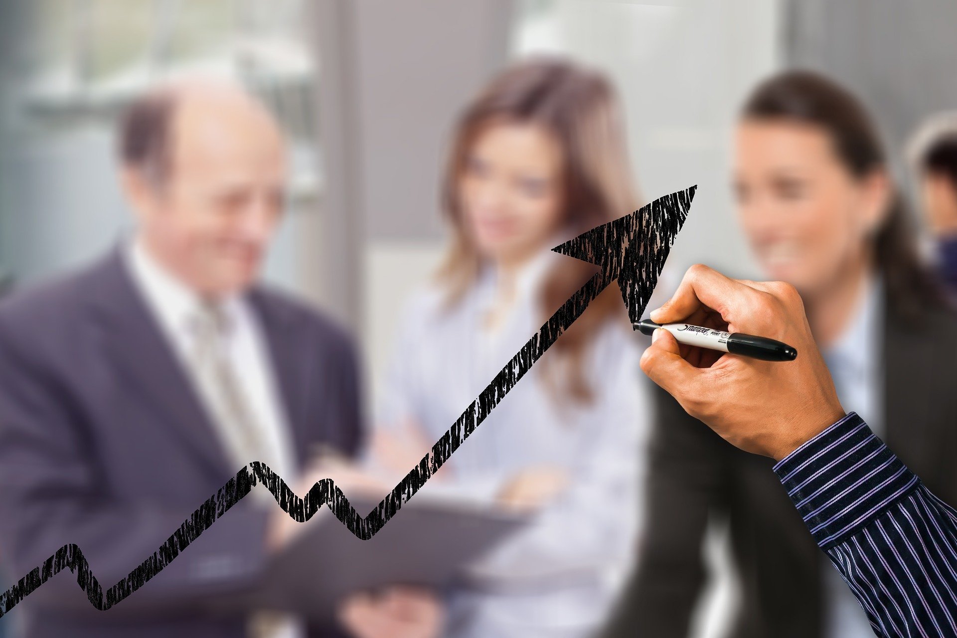 Image shows an upward trend arrow drawn on glass with a black marker. Behind the glass is three businesspeople smiling and looking at a document together.