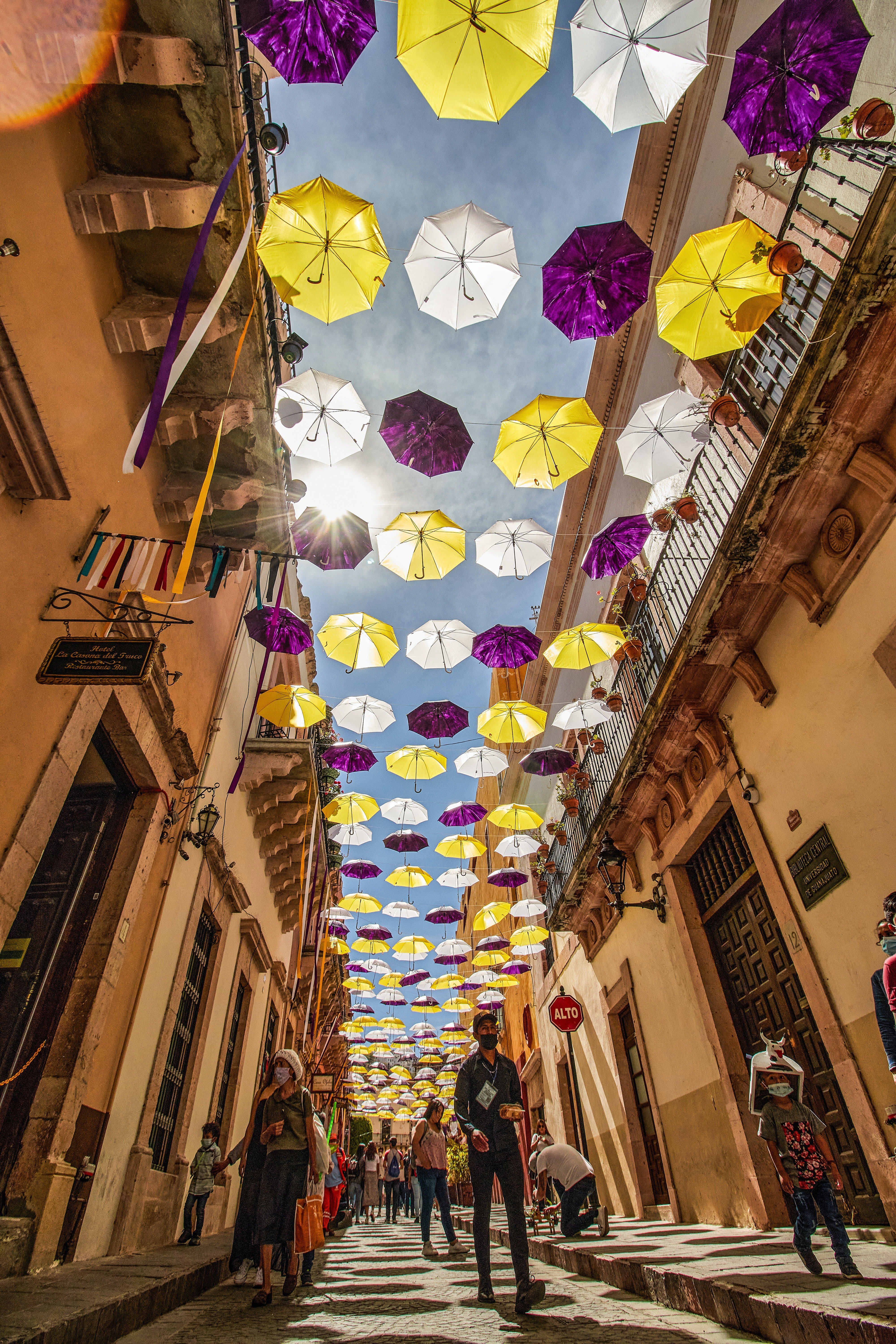 Image of a long walkway in between tall buildings. The image is looking up at white, purple, and yellow umbrellas strung across the walkway