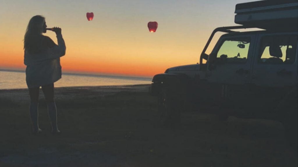 Frau steht neben einem Jeep Wrangler im Sonnenuntergang mit roten Himmelslaternen