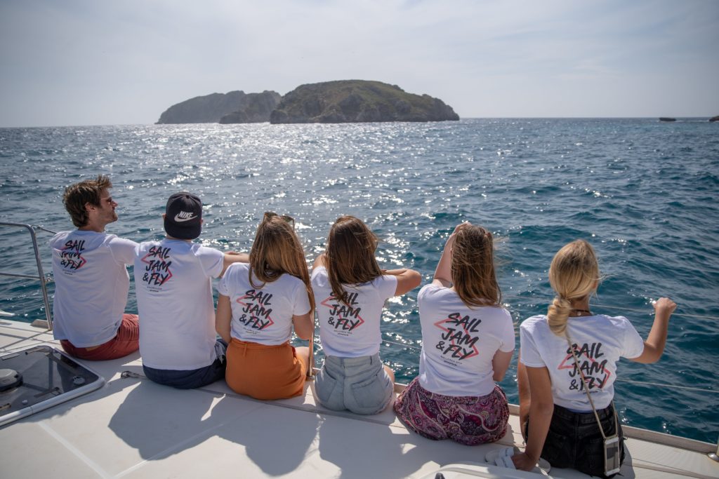 Crewmitglieder in Sail,Jam & Fly T-Shirts von hinten auf Rand des Bootes sitzend