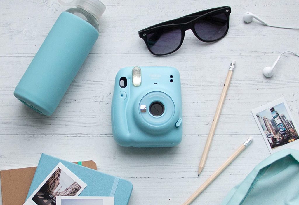 A blue polaroid camera laid on top of a white wooden table with other travel essential must haves on the table.