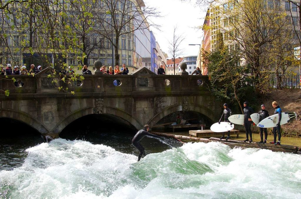 Die Eisbachwelle in der Stadt München ist bekannt als Riversurfing Spot in Deutschland, Es ist ein Surfer im Wasser und 4 Surfer warten am Wasserrand. Alle tragen schwarze Neoprenanzüge. 