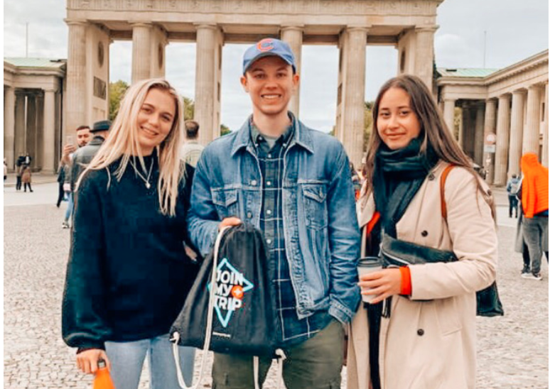 3 friends standing infront of Brandenburg gate holding a JoinMyTrip bag. 