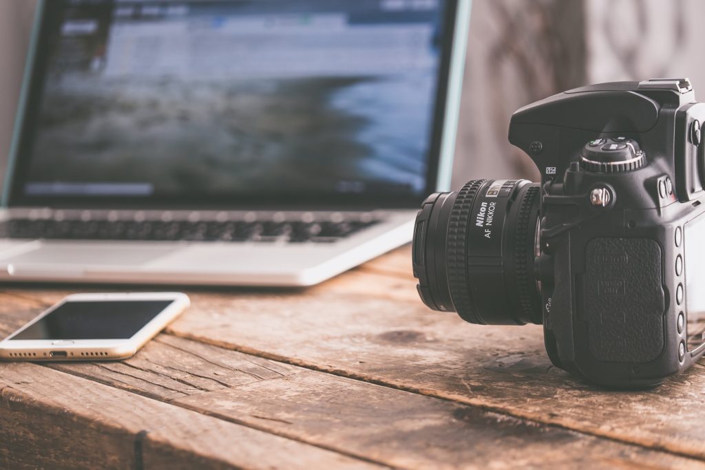 a camera and a phone placed on a wooden table with a laptop in the distance 