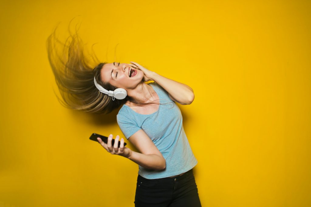 A girl singing infront of a yellow background with white headphones and black phone in her hand during the winter lockdown
