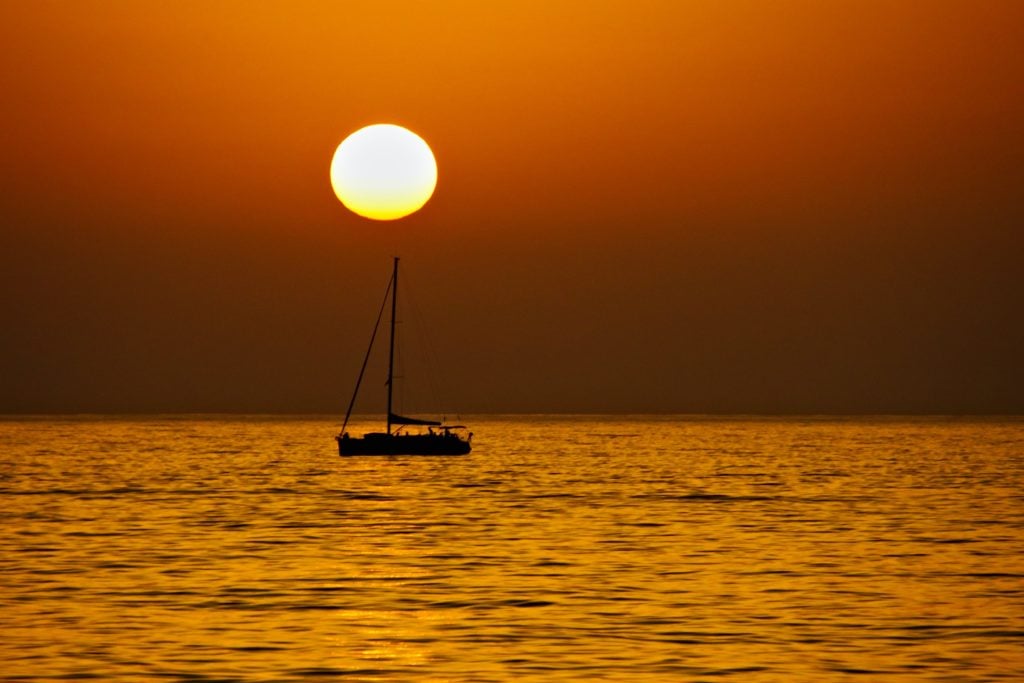 Ein kleines Segelboot fährt auf dem Meer und im Hintergrund sieht man die untergehende Sonne.