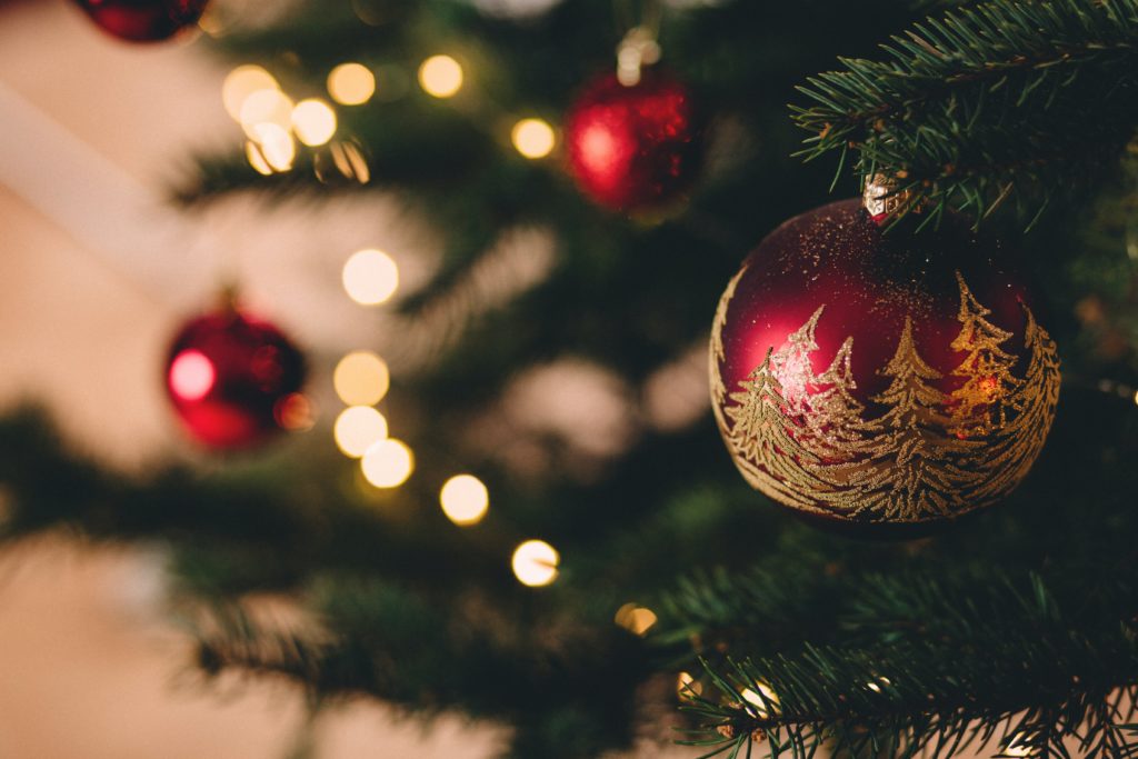 Eine rote Weihnachtskugel mit goldenen Weihnachtsbäumen drauf und im Hintergrund leuchtet eine Lichterkette 