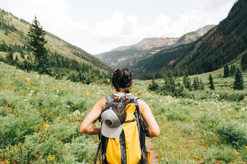 Frau mit gelbem Rucksack wandert in bergiger Umgebung, von hinten zu sehen 