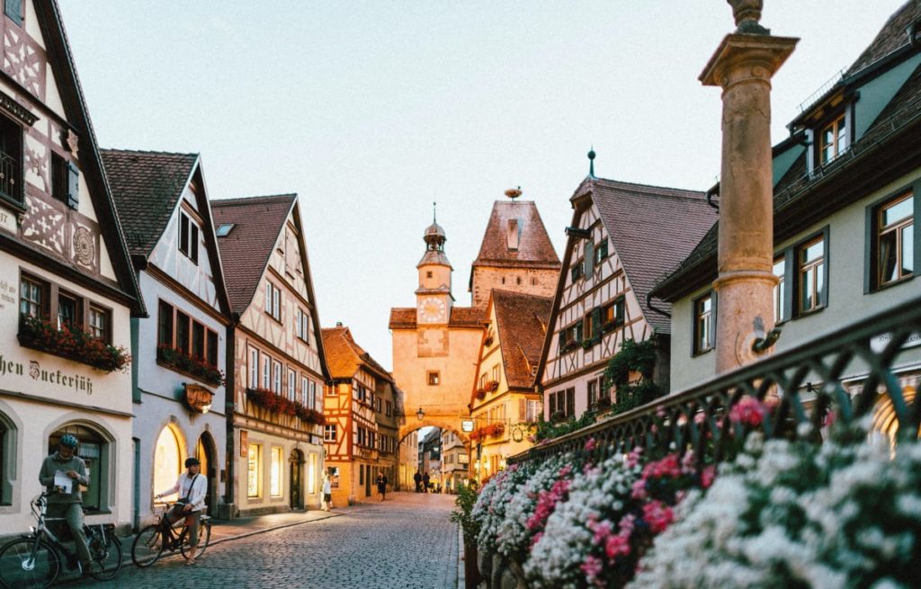 Rothenburg ob der Tauber ist ein malerisches Städtchen in Bayern. Zu sehen sind alte Fachwerkhäuser und spitze Kirchtürme im Sonnenuntergang und ein alter Brunnen mit Weißen und pinken Blumen 