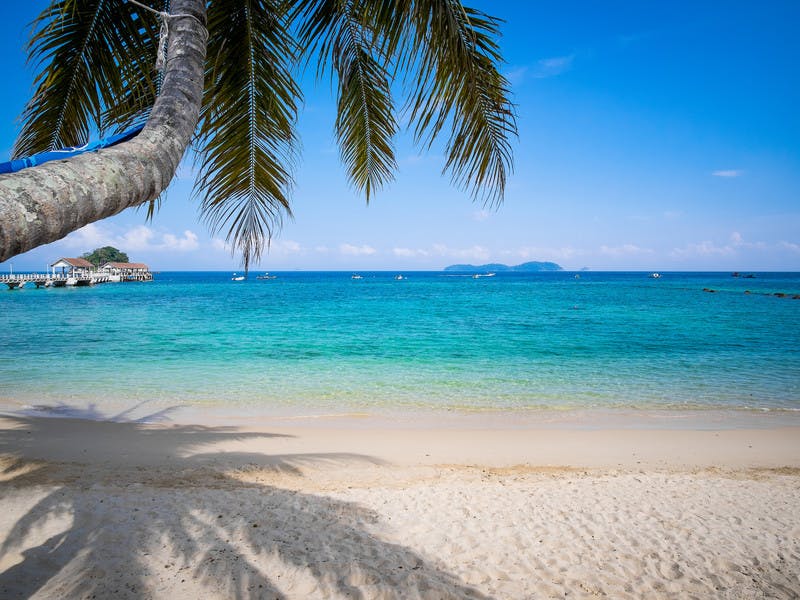 Strand von Tioman mit teilweise zu sehender Palme, weit im Hintergrund ein Steg und Land 