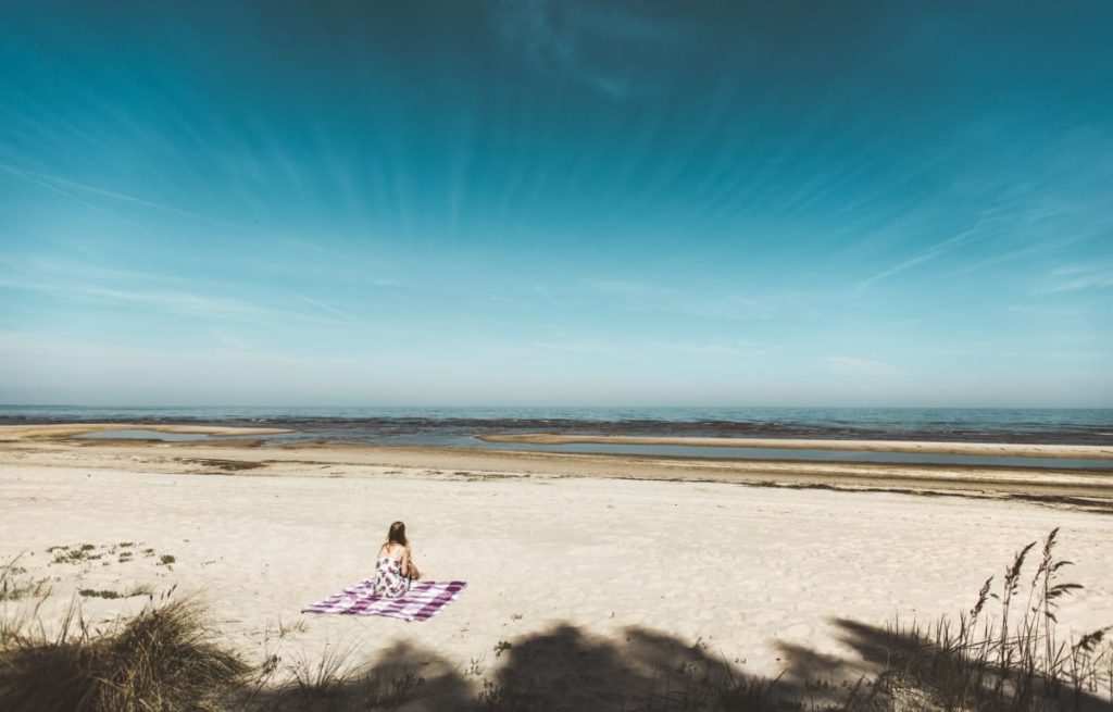Frau sitzt auf einem rot-weiß karierten Tuch am weiten Strand mit Schilf im Vordergrund und der Ostsee im Hintergrund. Darüber erstreckt sich sehr strahlend blaue Himmel 