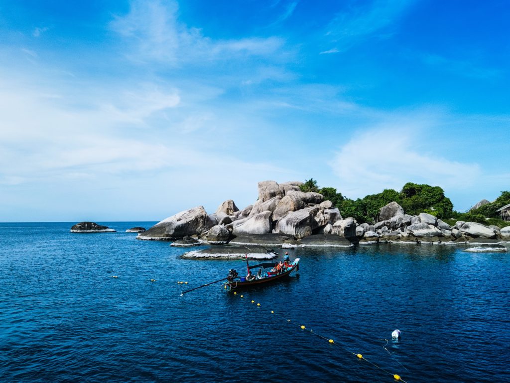 Eine kleine Insel auf großen Felsen grünen Bäumen und davor fährt ein kleines Boot auf dem klaren dunkelblauen Wasser