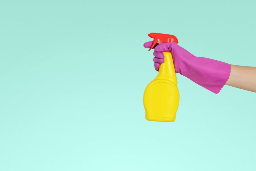 Hand mit einem pinken Reinigungshandschuh hält eine gelbe Sprühflasche mit rotem Kopf