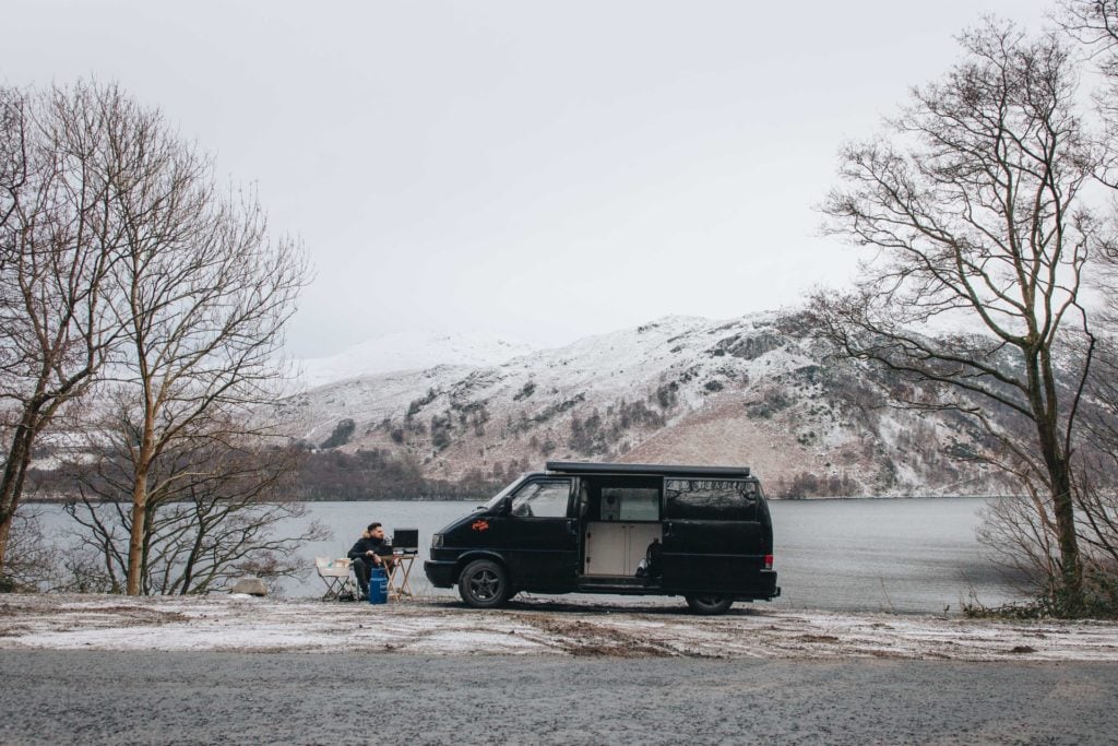 man sitting outsdie on a chair next to his black campervan whilst on a road trip in the snow 