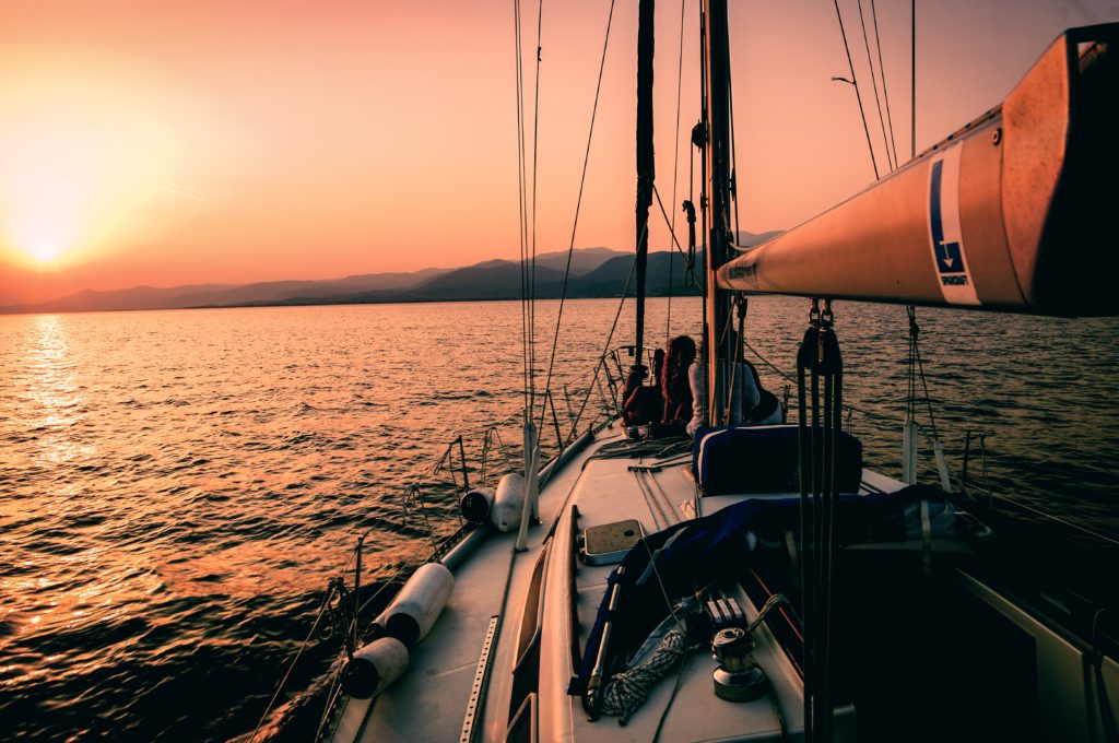 Man sieht die Spitze eines Segelbootes. Vorne sitzen Menschen und beobachten den rosa Sonnenuntergang