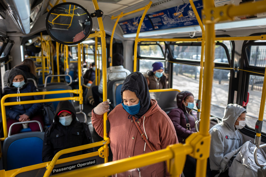 Ein Bus voller Menschen, die Masken tragen. Am Eingang steht eine Frau mit einer Kapuze, die auf den Boden schaut.