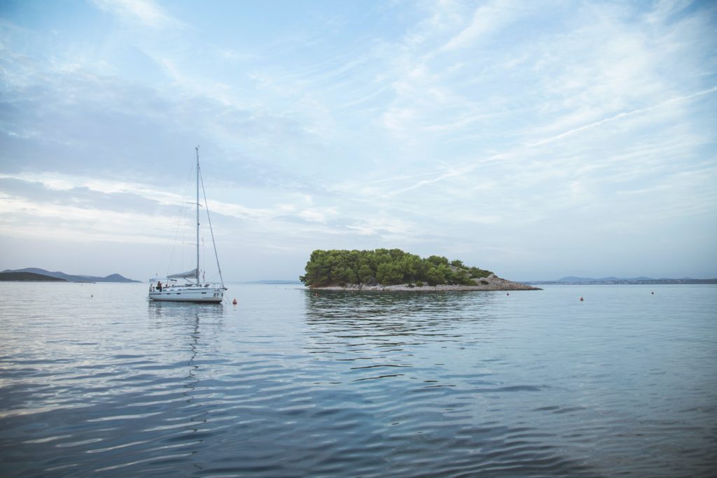 Ein einsames, weißes Segelboot fährt vor einer kleinen grünen Insel entland. Es ist umringt von blauem Wasser.