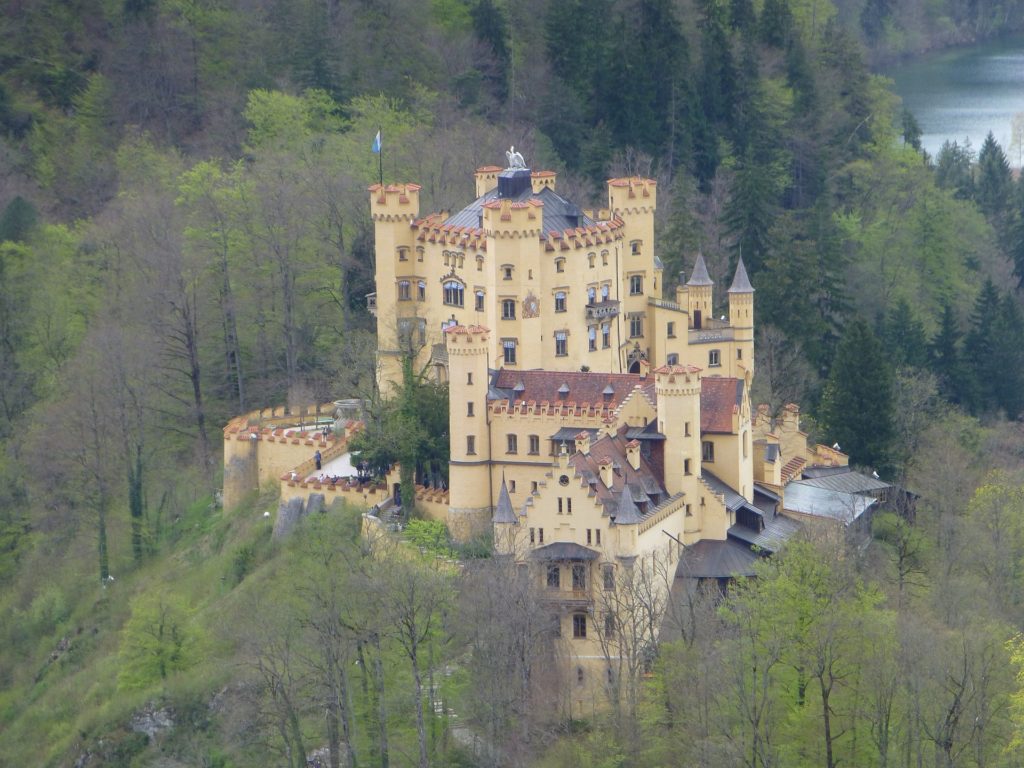 a yellow castle surrounded by a beautiful landscape of water and green trees. the Hohenschwangau is located in Bavaria