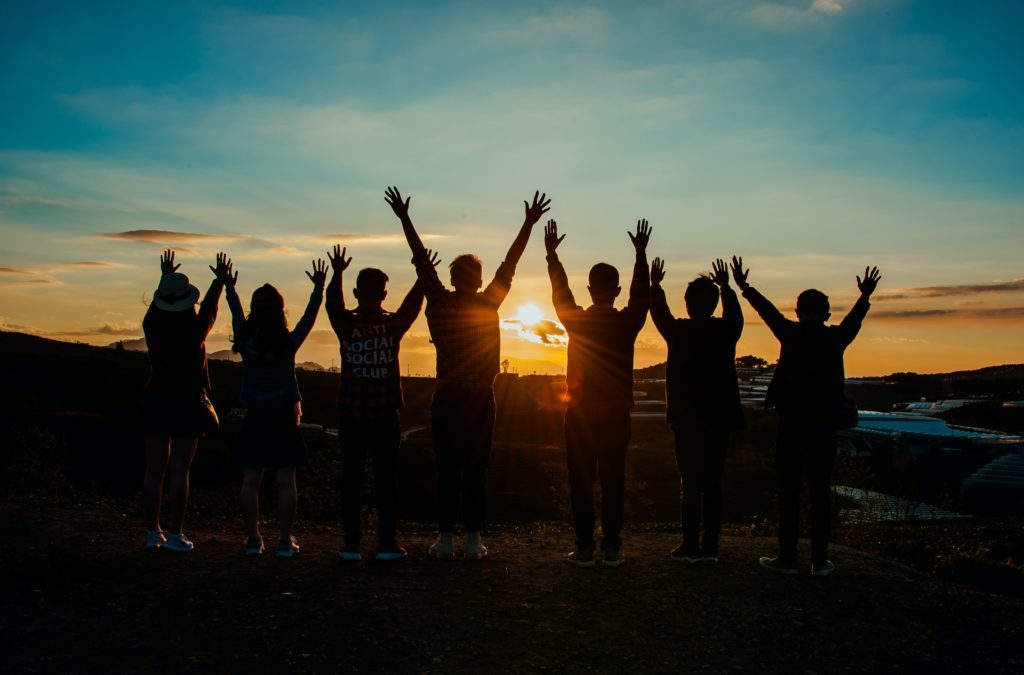 Ein Bild von Menschen, die während eines Sonnenuntergangs alle zusammen ihre Hände in die Luft strecken und dabei mit dem Rücken zur Kamera stehen.