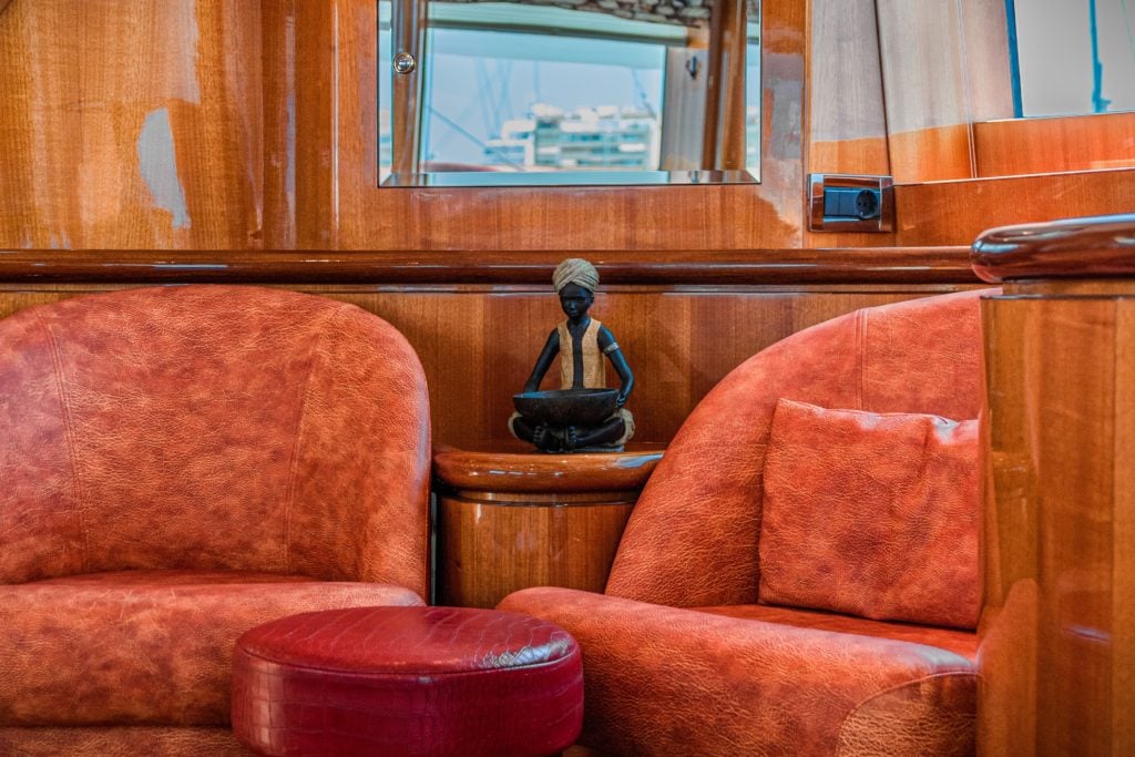 Innensicht Yacht mit 2 orangenen Sesseln und rotem Hocker, dazwischen eine kleine Menschenfigur. Restliche Ausstattung besteht aus Holz