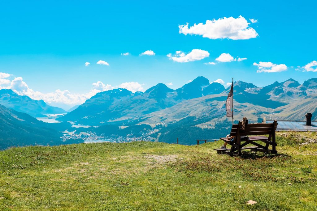 Muottas Muragl with a bench on the green grass where a hiker is resting and enjoying the view.