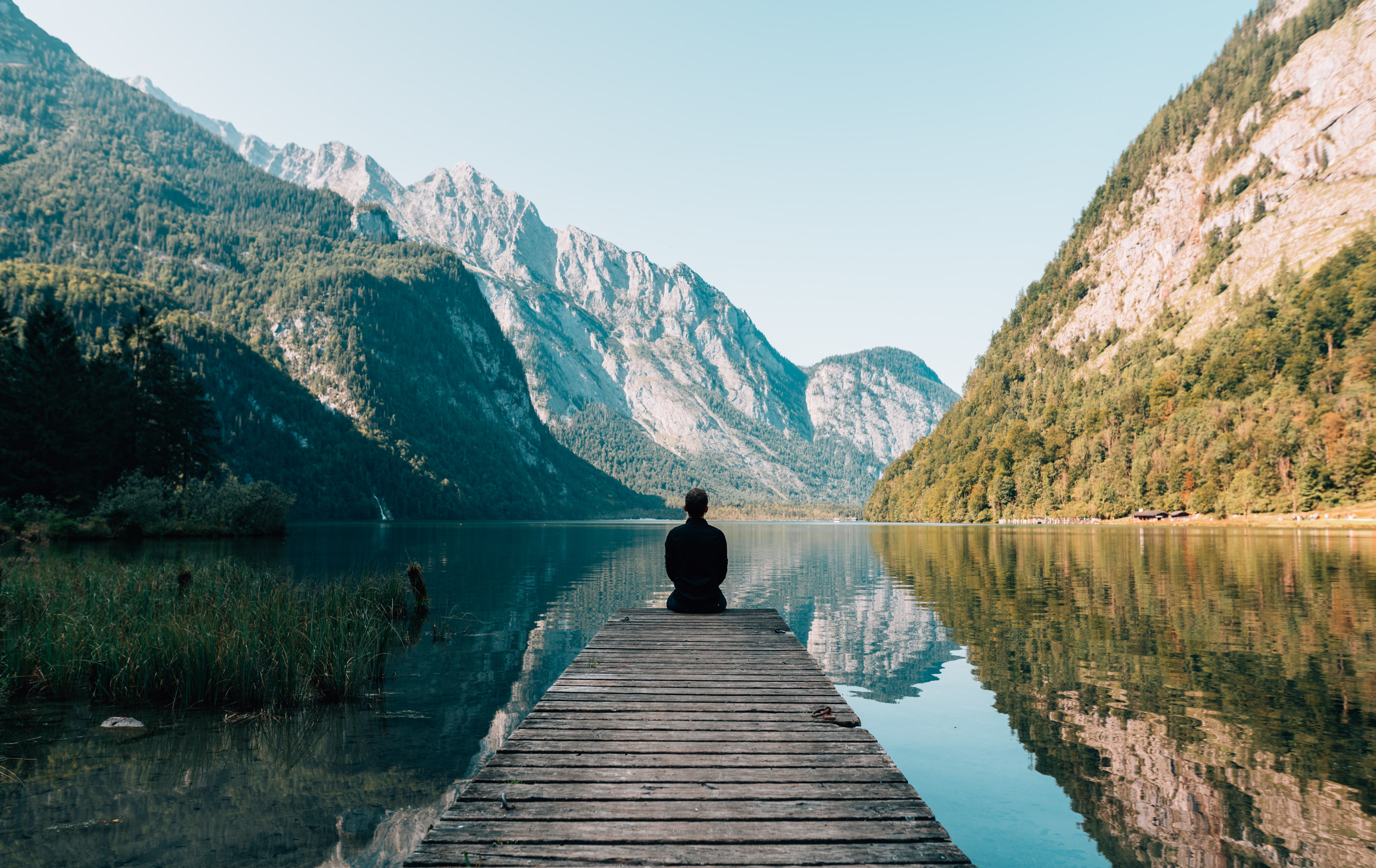 Eine Person sitzt auf einem Steg an einem See und Blick auf die Berge rundherum. 