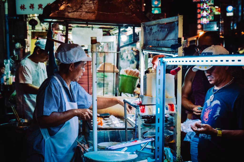 Mann mit Mütze kocht Streetfood an kleinem Stand, um ihn herum Kunden und Köche