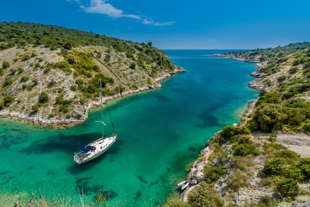 Ein Segelboot fährt durch eine Meerenge mit türkisblauem Wasser und grünem Festland.