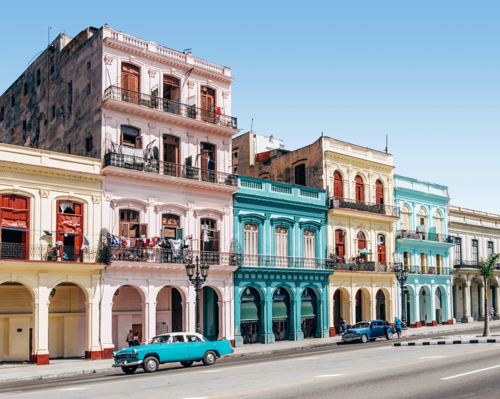 two blue cars sitting outside the vibrant and bright buildings in Cuba