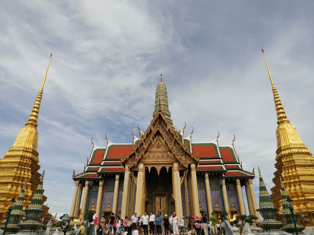 Blick auf den Wat Phra Kaew Tempel. Viele Menschen stehen vor dem Eingang, welcher umringt ist von 2 goldenen Türmen
