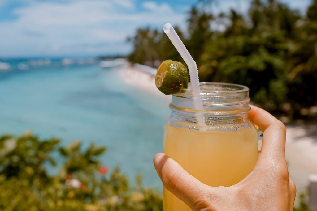 a drink being held by a hand with the view of a beach 
