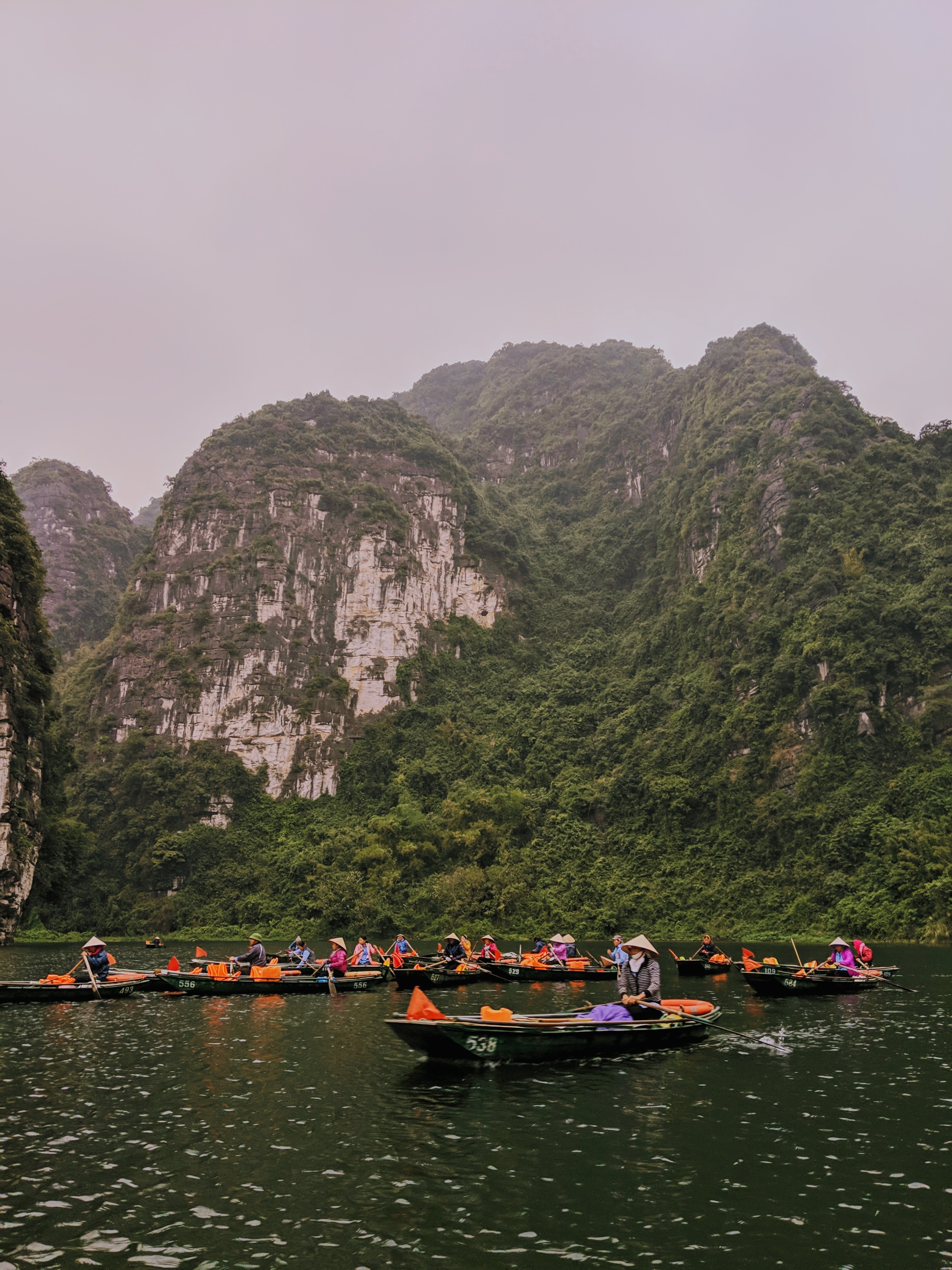 Eine Bootstour Trang An Vietnam. Im Hintergrund sieht man grün bewachsene Hänge und im Vordergrund sieht man mehrere typisch vietnamesische Boote mit bunten Stoffen. 