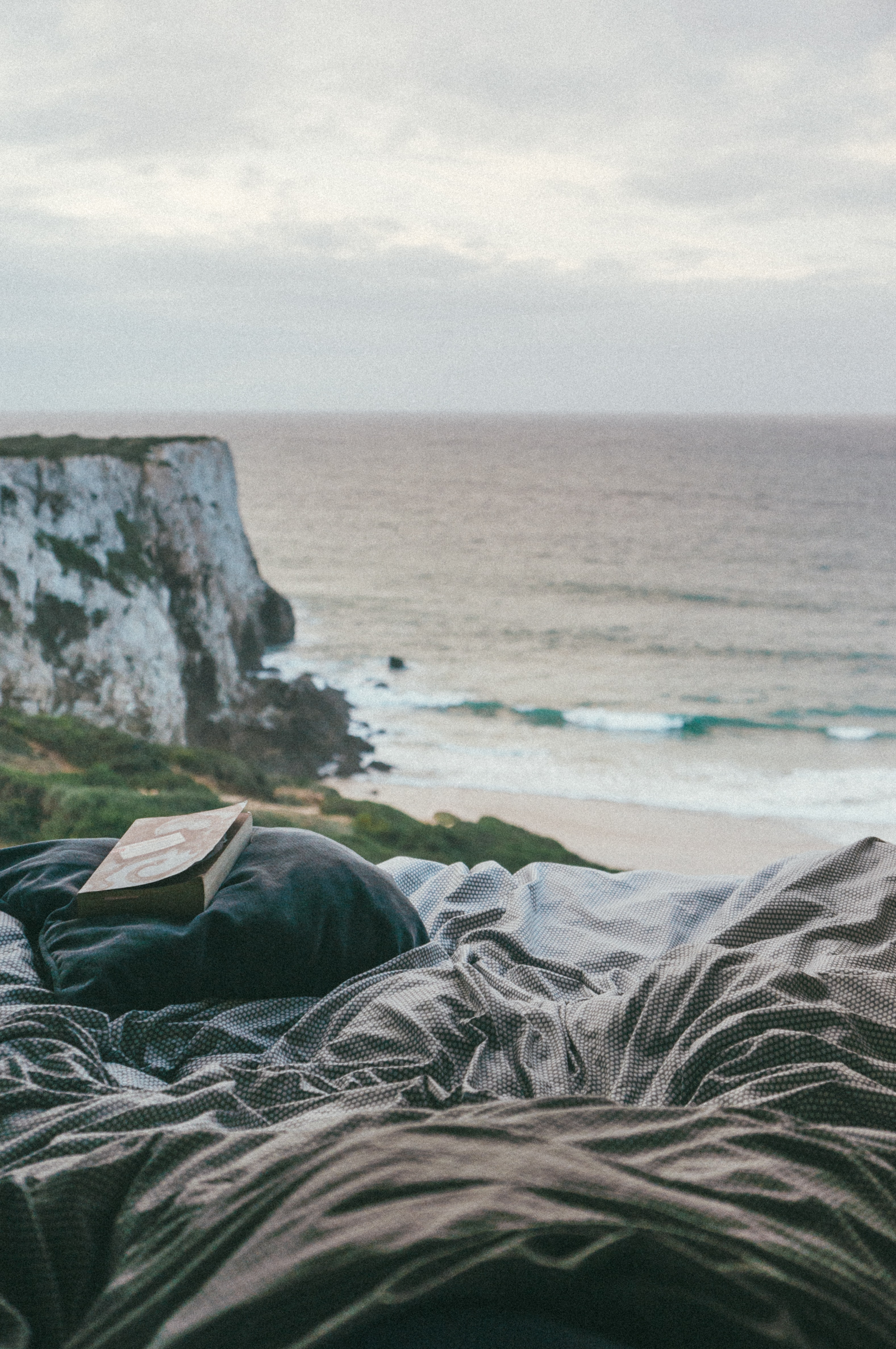 Ein gemütliches Nest aus Decken in denen eine lesende Person sitzt, direkt an den Klippen mit Blick über das Meer.